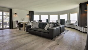 A modern living room with a grey sofa, a dining table, and a large window overlooking a scenic view. - House by the sea Comfortable Holiday Residence (Sylt)