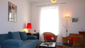 Un salon avec un canapé bleu, un fauteuil en osier avec des coussins rouges, une petite table et un lampadaire.