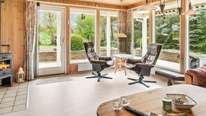 Una habitación acogedora con chimenea, dos sillones de cuero, una mesa de madera y ventanas grandes que dan a un jardín.