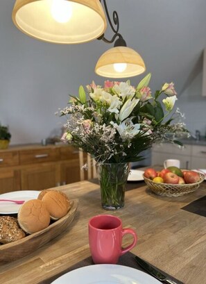 Un comptoir de cuisine orné d’un vase de fleurs, d’une tasse rose, d’un panier de pommes et d’une table en bois.
