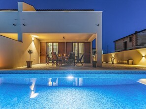 A modern outdoor pool area with a dining set and chairs, surrounded by a building with large windows and a tiled roof. - Family Villa With Pool, Wellness (Vodnjan)