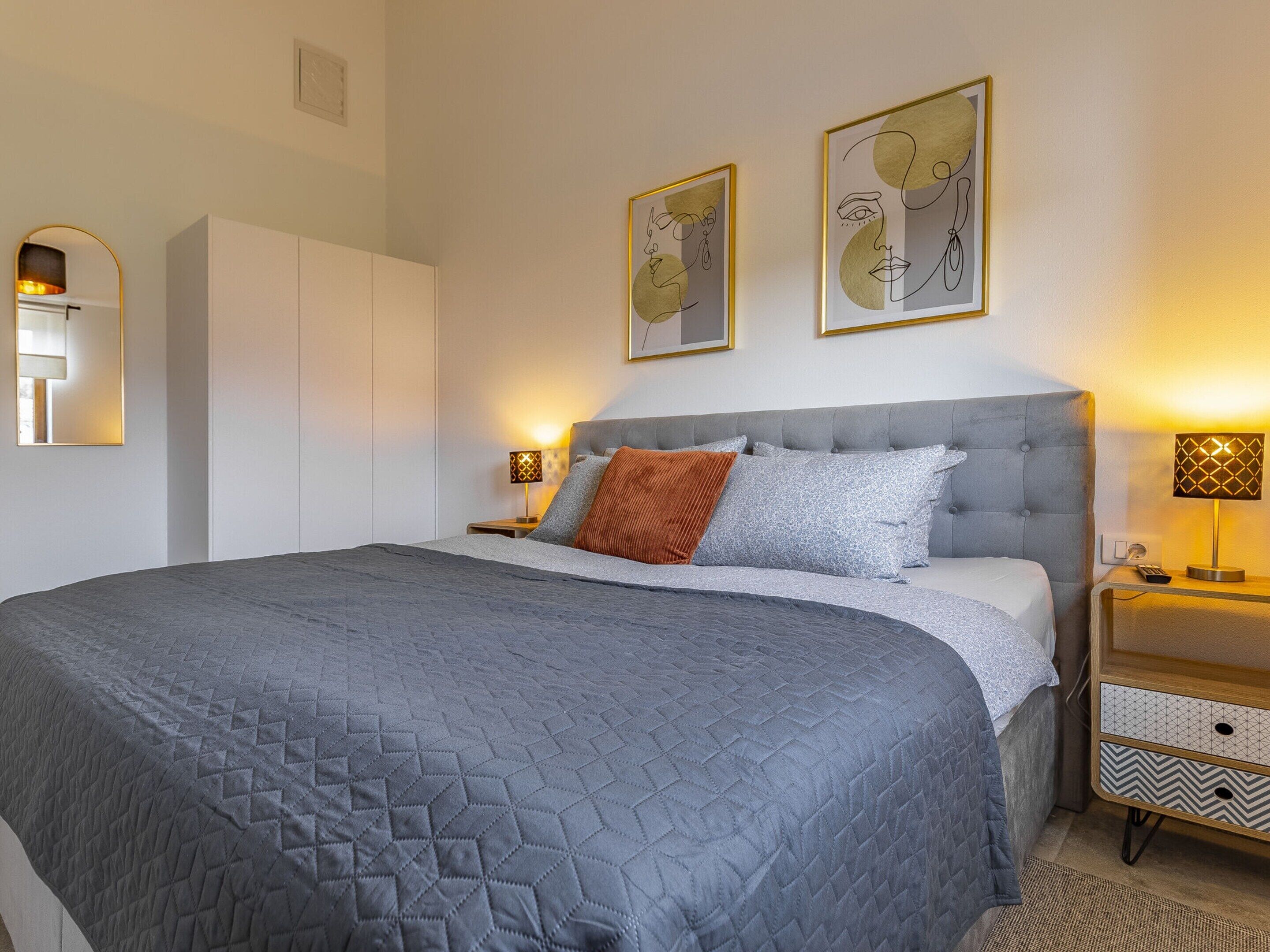 A neatly arranged bedroom with a large bed, a wardrobe, bedside lamps, and two framed artworks on the wall.