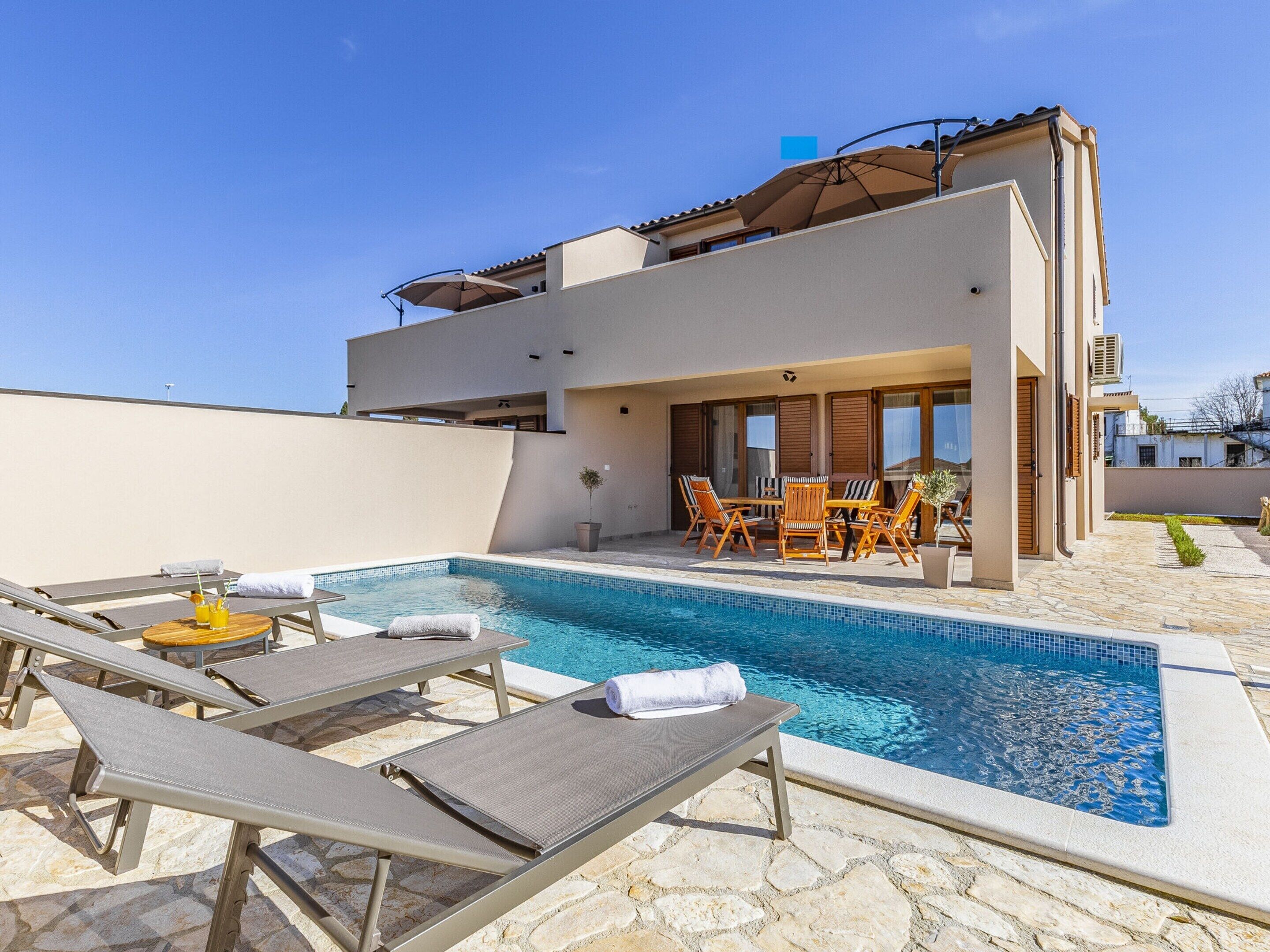 A modern pool area with lounge chairs and a table, a clear blue pool, and a building with large windows and a flat roof.