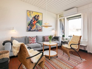 Un salon moderne avec un canapé gris, une table basse ronde, des fauteuils en osier, un tableau coloré accroché au mur et une grande fenêtre avec des rideaux.