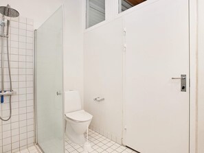 Une petite salle de bain blanche, équipée de toilettes, d’une douche avec un pommeau de douche amovible et d’une porte dotée d’une poignée.