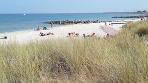 Ein Strand mit sonnenbadenden Personen, einem großen roten und weißen Streifen Sonnenschirm und einem Felssteg im Hintergrund.