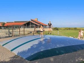 Un terrain de jeux avec un trampoline et des structures d’escalade, un bâtiment avec un toit rouge et, au fond, un champ d’herbe.