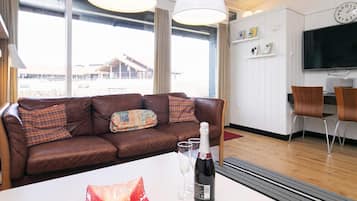 A modern living room with a brown leather sofa, a white table with a bottle and glasses, and a large window with a view of a building outside.