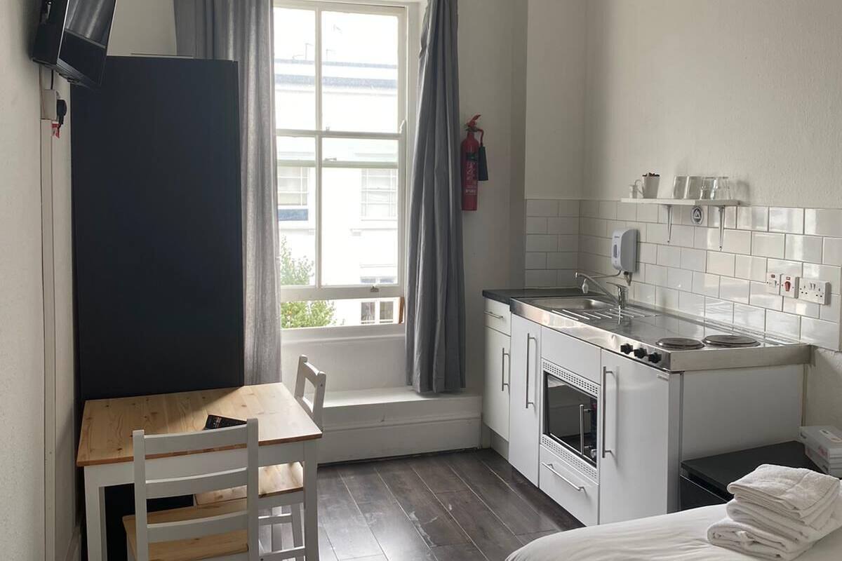 Une chambre d’hôtel de taille réduite comprenant un lit, une petite kitchenette, une table à manger avec des chaises, un téléviseur et une fenêtre avec des rideaux.