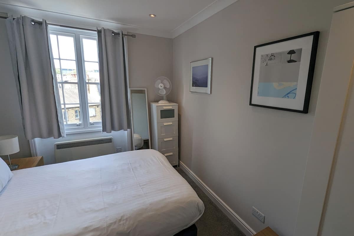 Une chambre d’hôtel comprenant un lit, une table de chevet avec une lampe, une commode, une fenêtre avec des rideaux et un tableau accroché au mur.