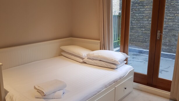 A neatly made bed with white linens and pillows, a white bedside table, and a view of a stone wall through a glass door. - The Beverley Apartments (London)