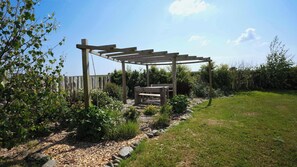 A wooden pergola structure in a garden with a bench underneath, surrounded by various plants and a grassy area.