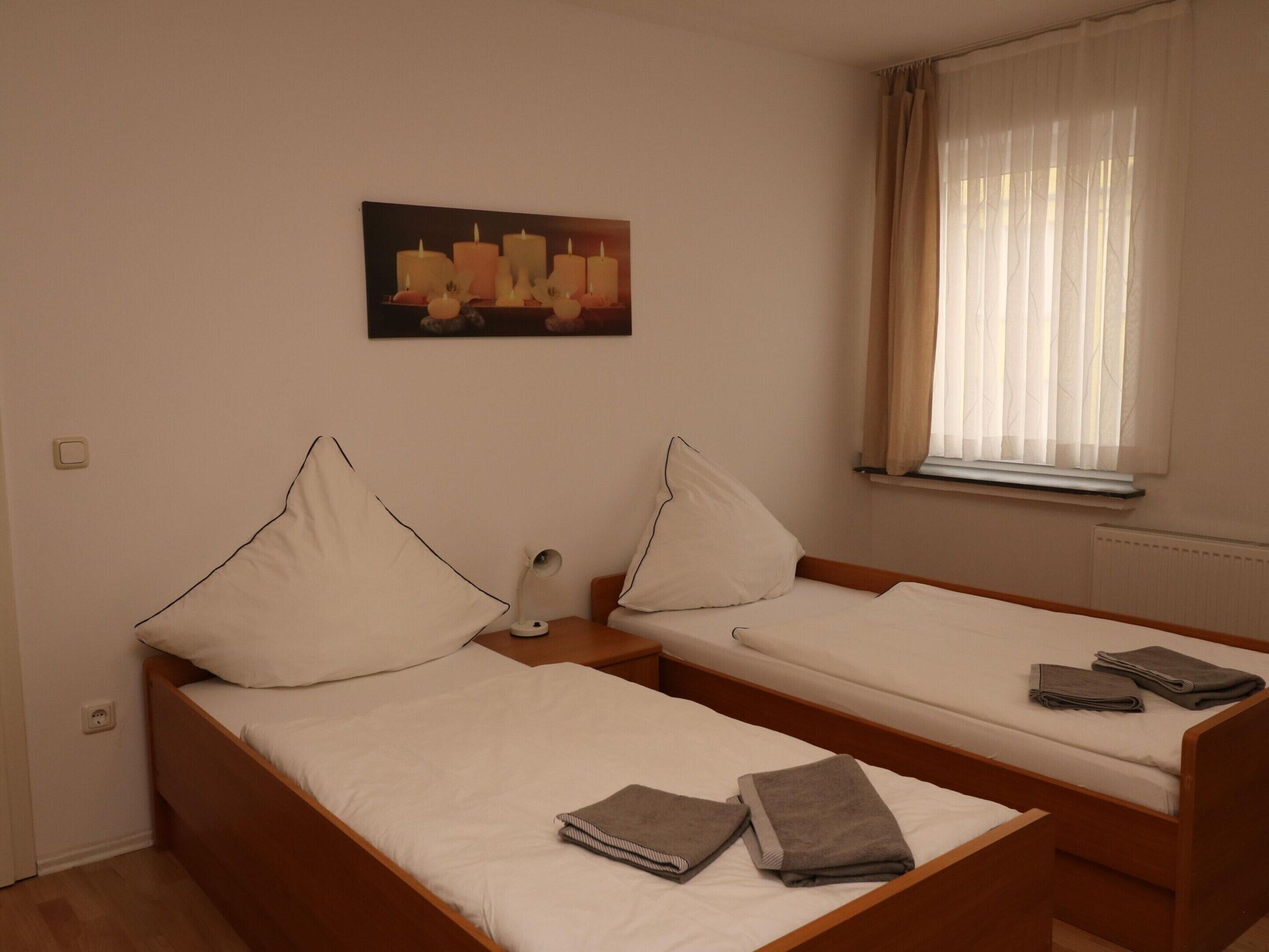 Una habitación con dos camas individuales, una mesita de noche con lámpara, un cuadro en la pared y una ventana con cortinas.