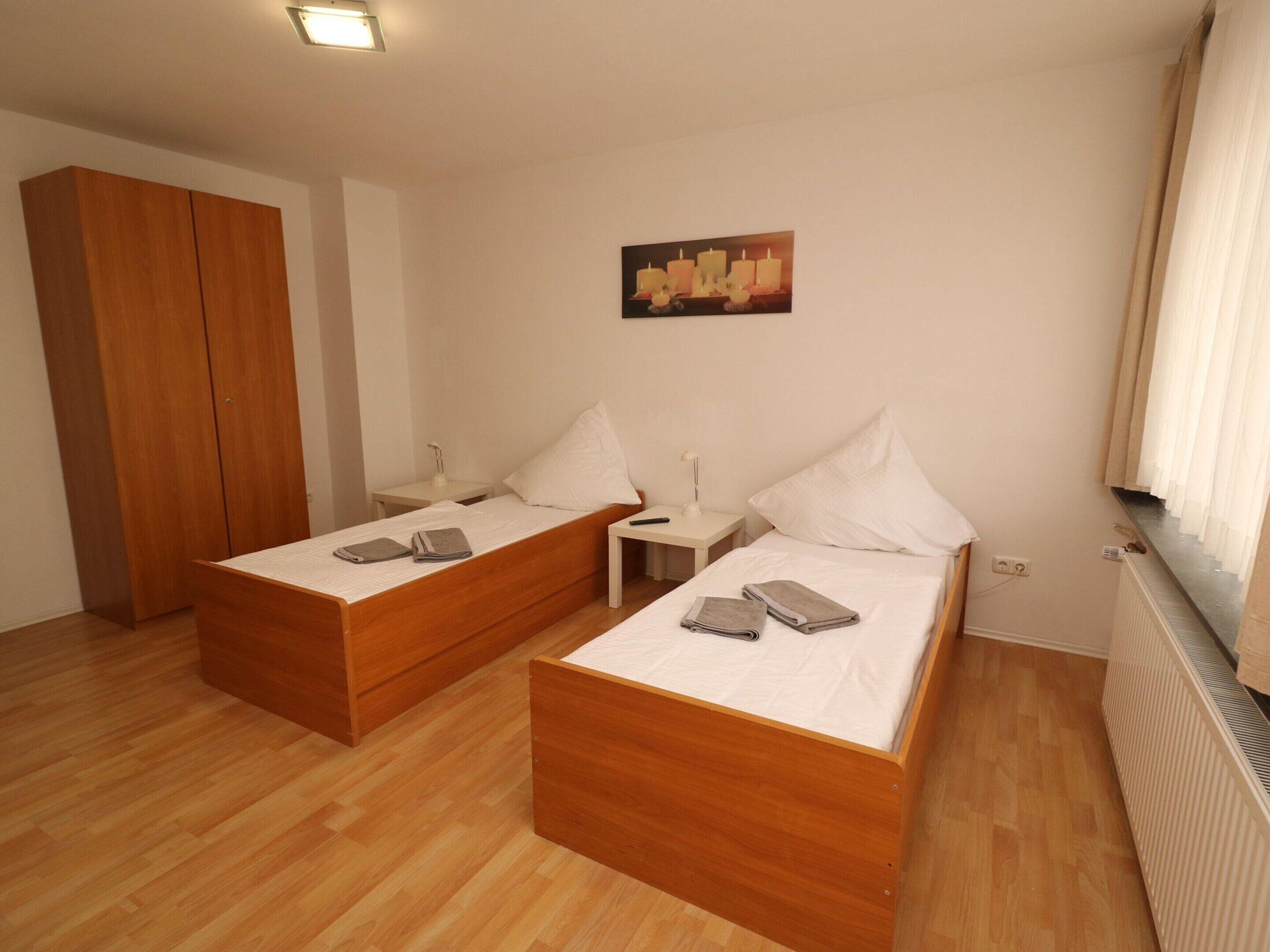 Un dormitorio con dos camas de madera, una mesita de noche con lámpara, un armario de madera y un cuadro en la pared.