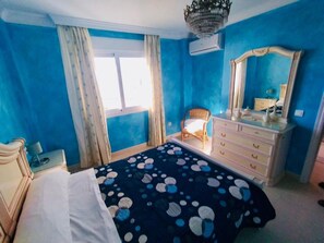 Relax in this serene bedroom with a large bed, dresser, and wicker chair.