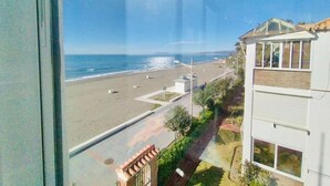 Uitzicht op een strand vanuit een raam, met een heldere lucht en een gebouw met balkon aan de rechterkant.