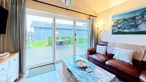 A living room with a brown leather sofa, a coffee table with a bowl of snacks, a painting of a coastal scene, and a sliding glass door leading to a backyard. - Stunning Coastal Beach House - Sleeping 4 in 2 Bedrooms - Pool and Beach Access Included. (Filey)