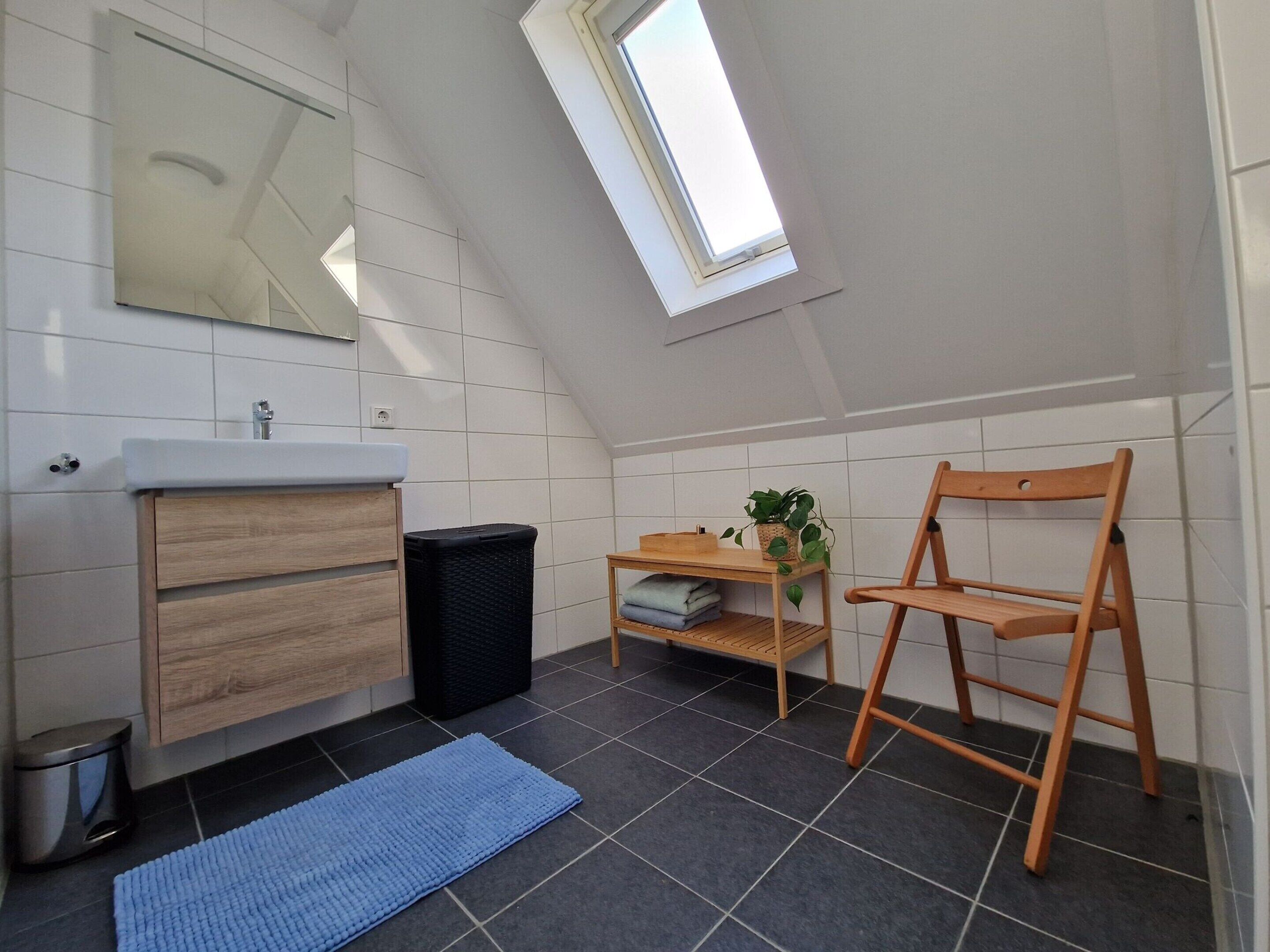 Un bagno moderno con una finestra a lucernario, un mobile bagno in legno, un cestino nero, un tappeto blu e una sedia in legno.