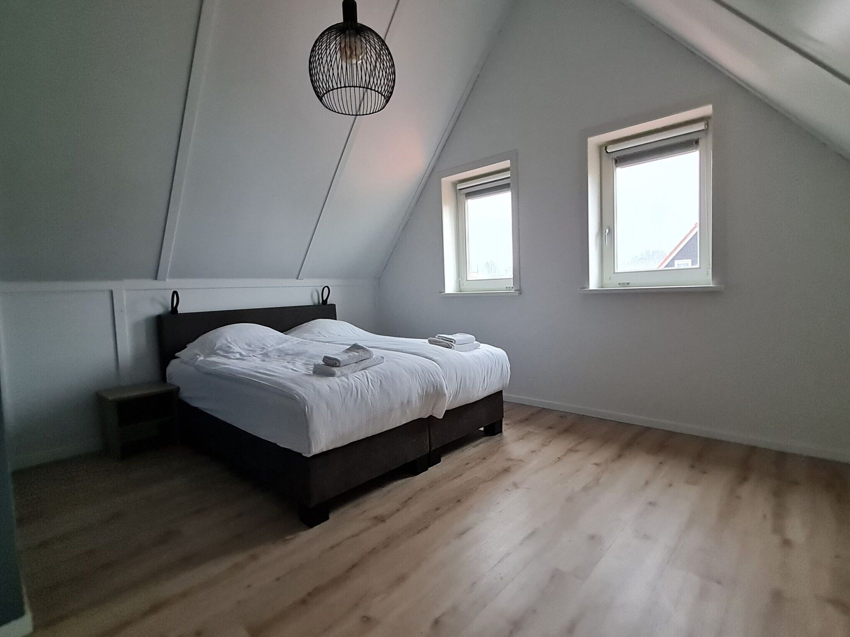 A bedroom with a sloped ceiling, a bed with white linens, two windows, and a hanging light fixture.