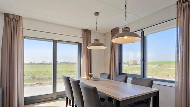 A modern dining area with a large table and chairs, large windows with curtains, and hanging pendant lights. - Holiday Home in Scherpenisse Near the Sea (Scherpenisse)