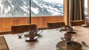 Ein Esstisch ist gedeckt mit Tellern, Tassen und Besteck, mit Blick auf verschneite Berge durch große Fenster.