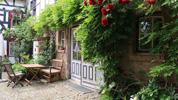 A courtyard with a cobblestone floor, a wooden table and chairs, and a brick wall covered in climbing plants and red flowers. - Apartment in Geiersthal (Weilmünster)