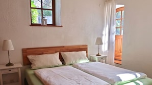 A double bed with a wooden headboard, two bedside tables with lamps, a window with curtains, and a view of greenery outside. - Apartment in Geiersthal (Weilmünster)