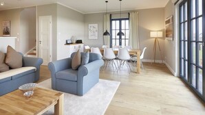 A modern living room with a sofa, a dining table with chairs, and a large window with curtains. - Luxury Roof Villa With Sauna and Fireplace (Dranske)