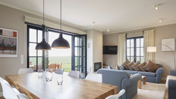 A spacious living room with a wooden dining table, white chairs, a blue sofa, and a large window offering a view of the outdoors. - Luxury Roof Villa With Sauna and Fireplace (Dranske)