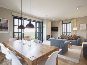 A spacious living room with a wooden dining table, white chairs, a blue sofa, and a large window offering a view of the outdoors. - Luxury Roof Villa With Sauna and Fireplace (Dranske)