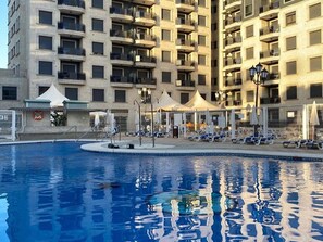 A swimming pool with lounge chairs and umbrellas, surrounded by a high-rise building with balconies. - Belvilla by OYO Apartment in Fuengirola (Fuengirola)