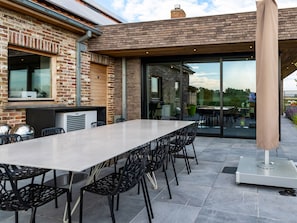 An outdoor patio with a long table and chairs, a brick wall, and a glass door leading to a garden. - Holiday Home in Haringe for ten Person (Poperinge)