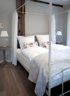 Un letto a baldacchino con lenzuola bianche e cuscini floreali, un comodino con una lampada e un pavimento in legno.