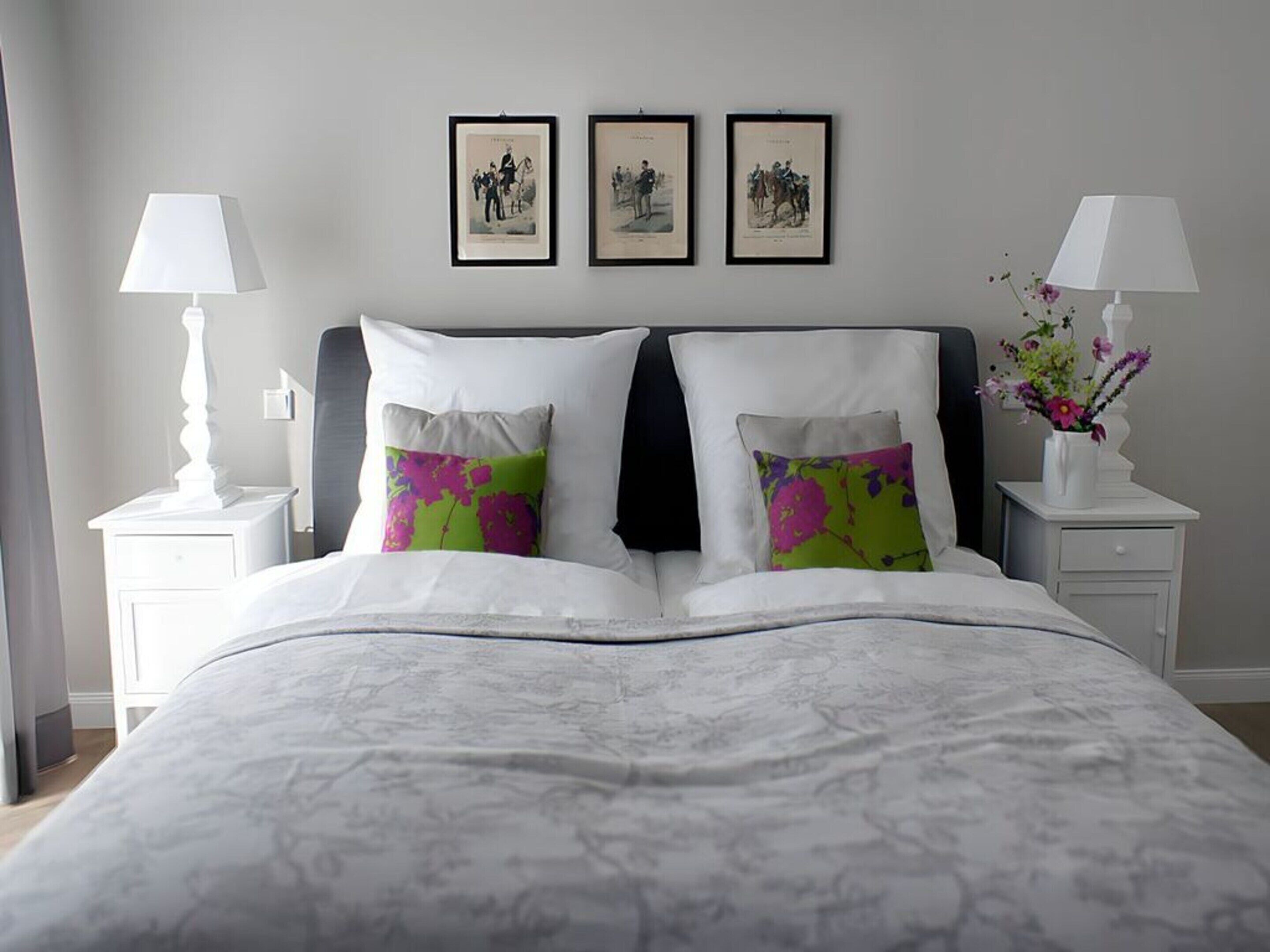 A neatly made bed with a white and gray patterned bedspread, two pillows with colorful floral designs, and a headboard. There are three framed pictures on the wall above the bed, and two bedside tables with lamps and a vase of flowers.