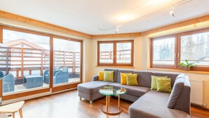 A modern living room with a grey sofa, yellow cushions, a glass coffee table, and wooden flooring. Large windows provide natural light and a view of the outside. - Nice Apartment in Langenfeld With Sauna (Laengenfeld)
