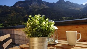 Uma mesa de madeira com um balde de metal cheio de plantas verdes, uma vela e uma caneca branca. Uma vista de montanhas é visível ao fundo.