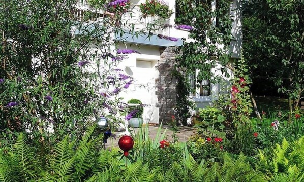 Un jardí amb plantes i flors diverses, una paret de pedra i un edifici amb façana blanca.