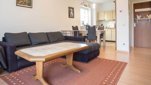 A living room with a sofa, a wooden coffee table, and a rug. There is a kitchen in the background with cabinets and a dining area.