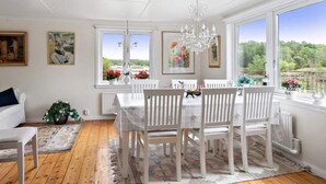 A dining area with a white table and chairs, a chandelier, and large windows with a view of greenery. - 5 Star Holiday Home in Stromstad (Stromstad)