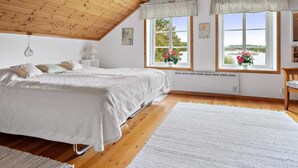 A bedroom with a sloped ceiling, wooden flooring, a bed with white bedding, two windows with curtains, and a view of greenery outside. - 5 Star Holiday Home in Stromstad (Stromstad)