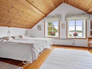 Quarto com teto inclinado, piso de madeira, cama com roupa de cama branca, duas janelas com cortinas e vista para o verde lá fora.