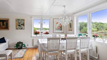 Uma sala de jantar com mesa e cadeiras brancas, um candelabro e grandes janelas com vista para vegetação.