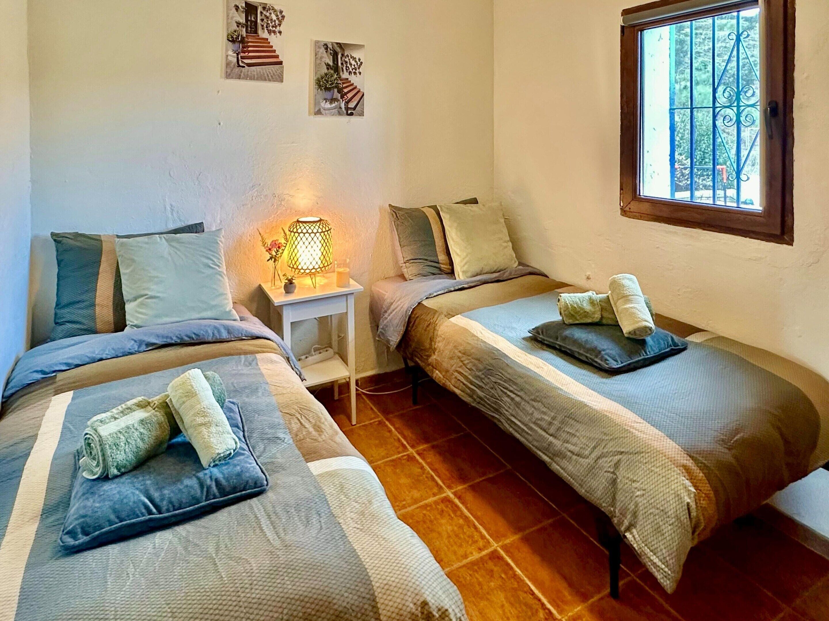 A bedroom with two single beds, a bedside table with a lamp, and a window with a view of trees.