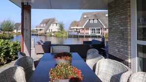 A covered outdoor seating area with a table and chairs, overlooking a canal with houses in the background. - Villa in Friesland With Sauna and Jetty (Delfstrahuizen)