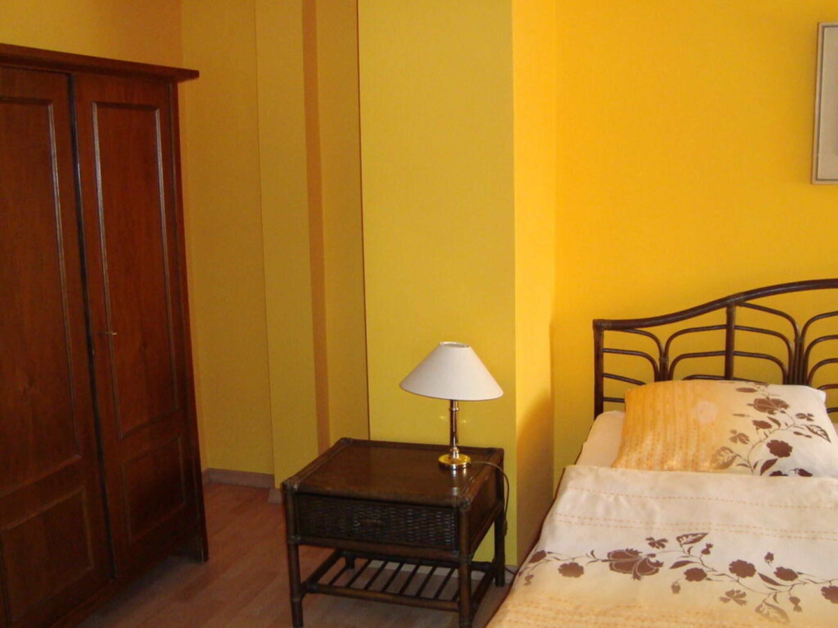 A bedroom with a wooden wardrobe, a small bedside table with a lamp, and a bed with floral bedding.