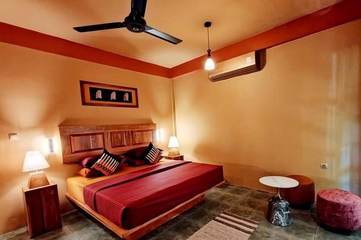A bedroom with a wooden bed, two bedside lamps, a ceiling fan, a small round table, and a red ottoman.