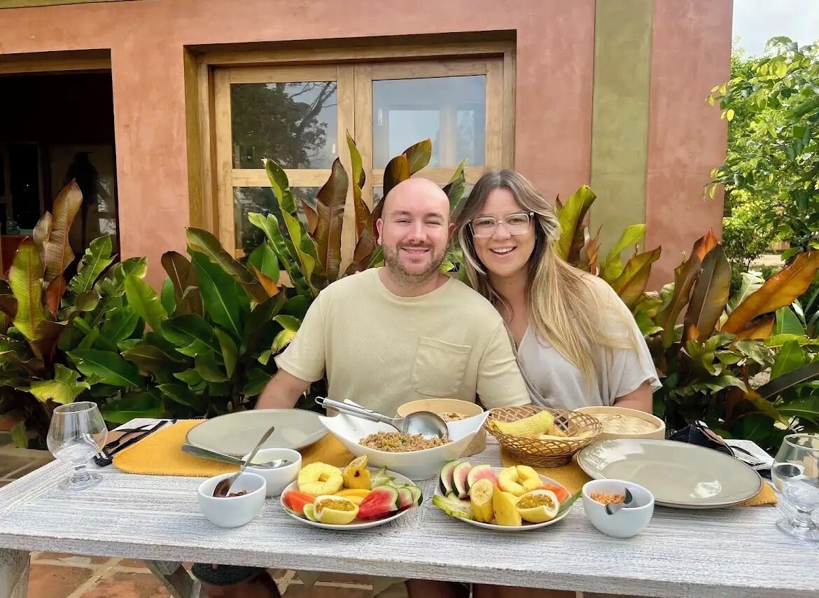 Ein Esstisch, reichhaltig gedeckt mit Frühstücksleckereien, darunter Früchte und Getreide, draußen inmitten üppiger Pflanzen mit einem Gebäude im Hintergrund.