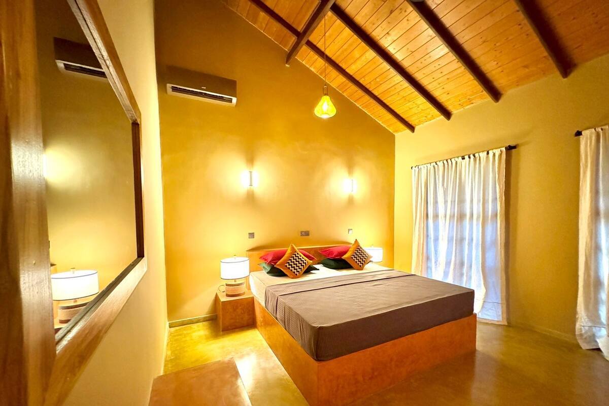 A bedroom with a wooden ceiling, a bed with pillows, a nightstand with a lamp, and a window with curtains.