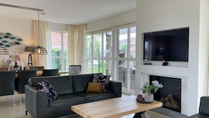 A modern living room with a sofa, a coffee table, and a fireplace. There is a TV mounted on the wall and a dining area with chairs and a table. - Beautiful Villa With Wellness (Delfstrahuizen)