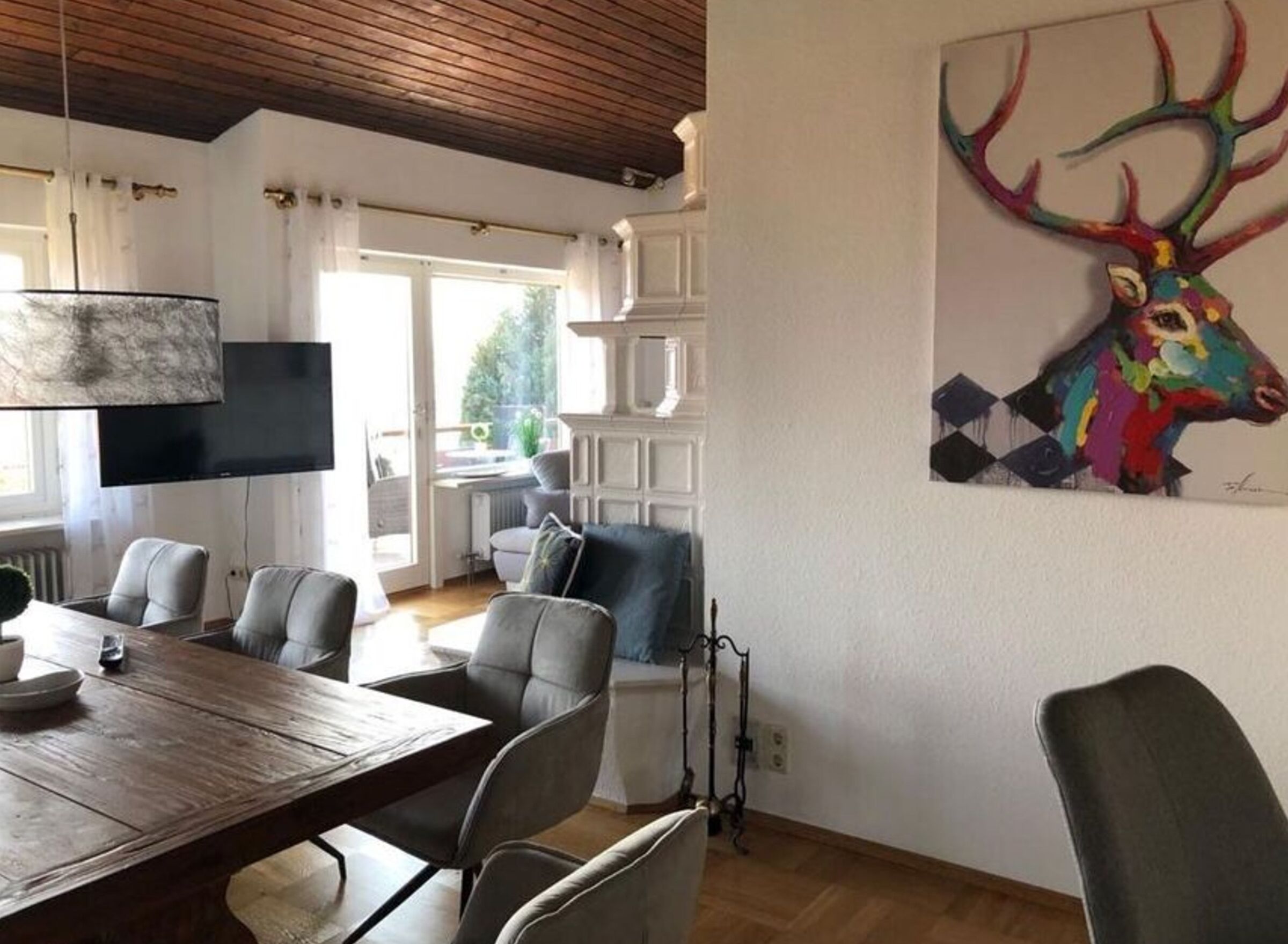 Uma sala de jantar com uma mesa e cadeiras de madeira, uma televisão fixada na parede e um quadro colorido de um veado na parede.